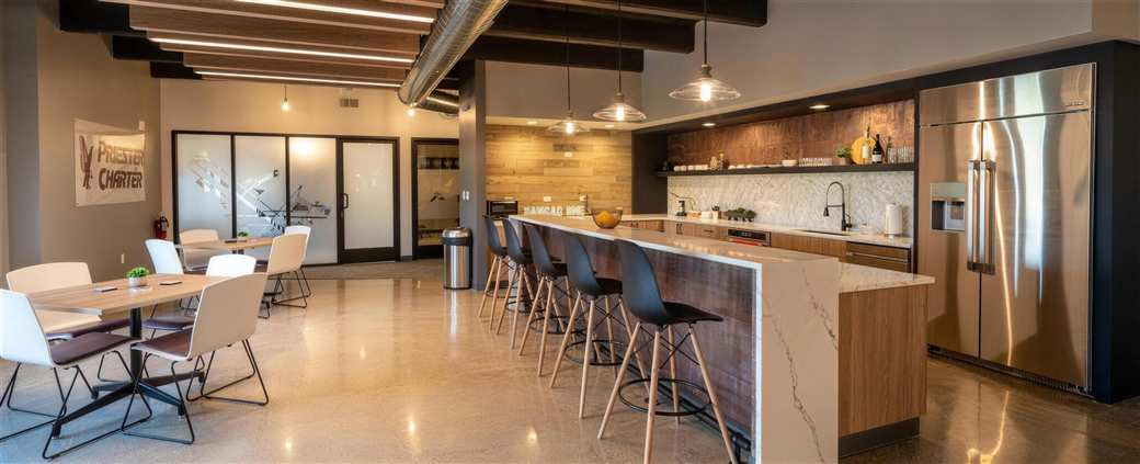 Priester Aviation crew and employee lounge completed by B&C, showing etched storefront glazing, a reclaimed wood feature wall with custom neon signage, a waterfall-edge quartz service island, and polished concrete flooring